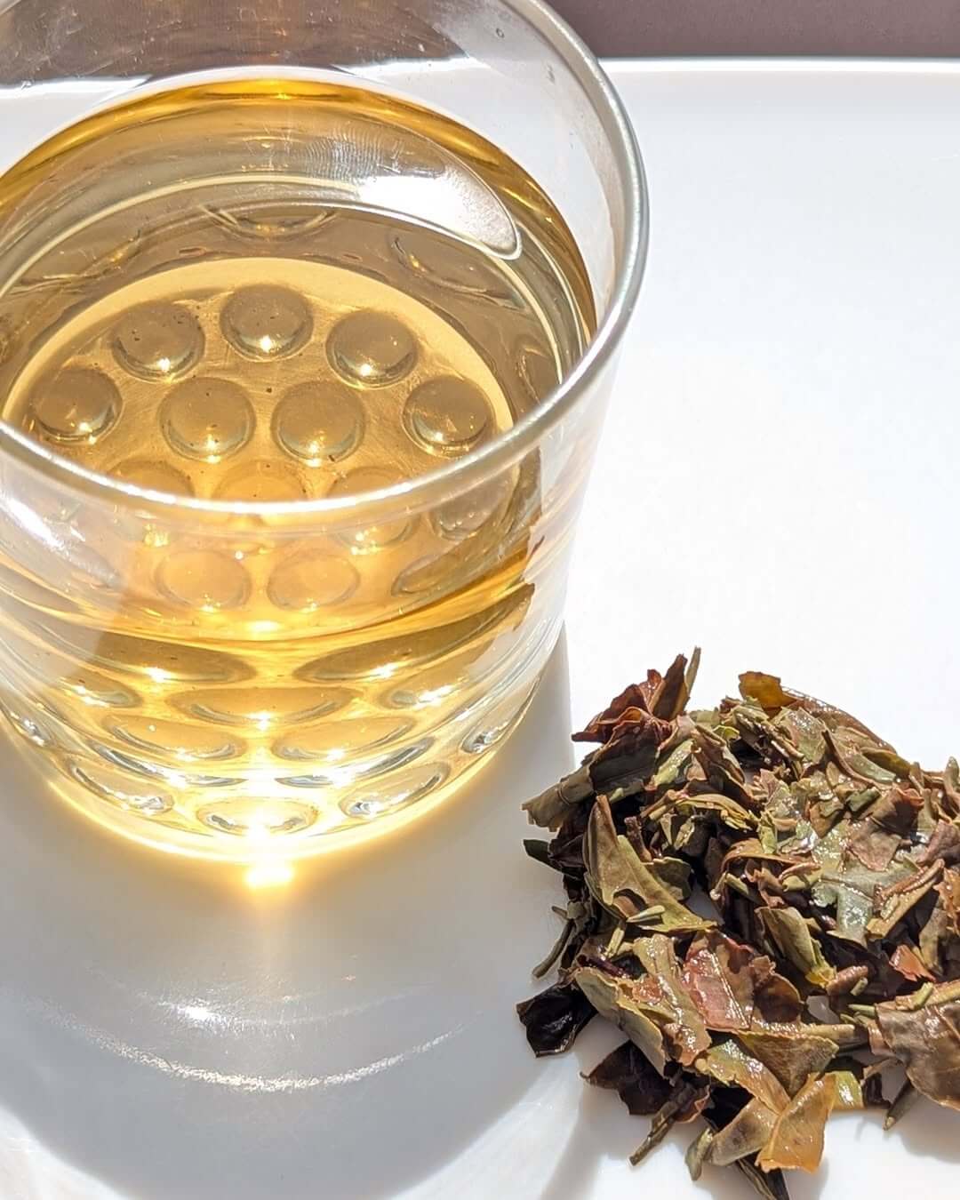 A clear glass of Aambari AV2 organic black tea next to loose leaves on a white surface.